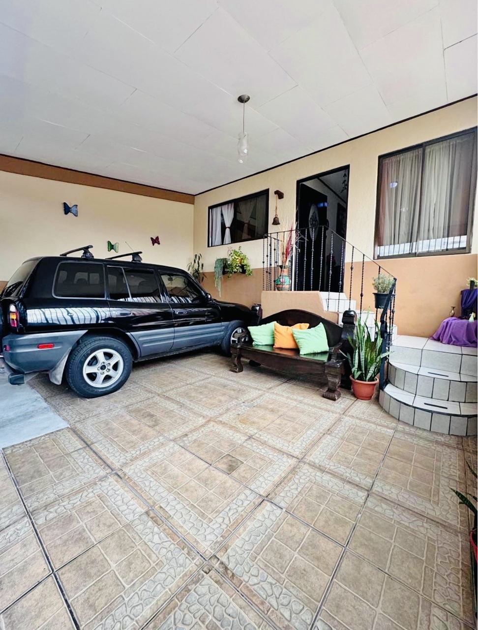 Patio frontal techado con baldosas de cerámica, vehículo estacionado y banco de madera — casa en-venta en Heredia